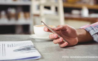 Man sitzt mit Kaffee und einer Zeitung am Tisch und liest etwas auf seinem Smartphone